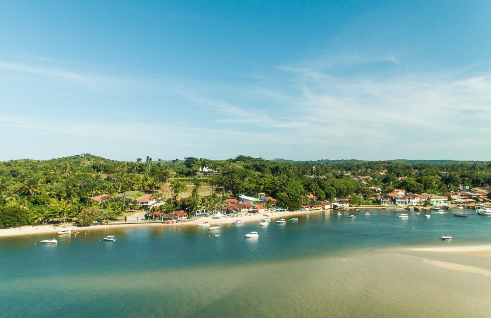 Vista aérea da Praia da Boca da Barra, com muitas árvores, barcos na água esverdeada e quiosques à beira-mar.