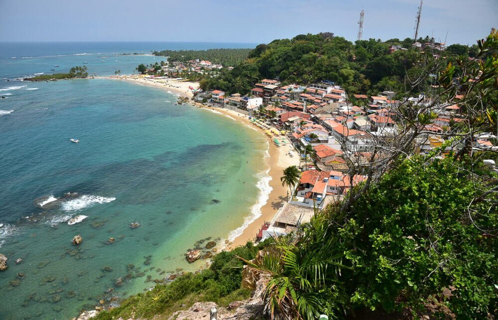 Vista aérea para praia em Morro de São Paulo, com águas esverdeadas, curta faixa de areia e várias casas e árvores à beira-mar.