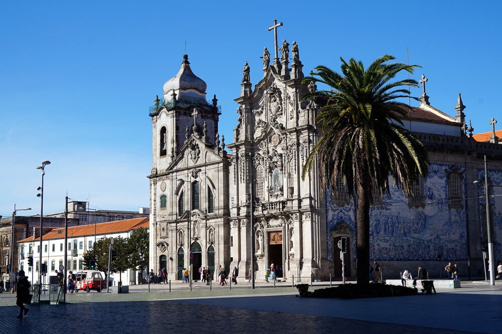 Pátio em frente à Igreja do Carmo, com coqueiros, postes e algumas pessoas caminhando.