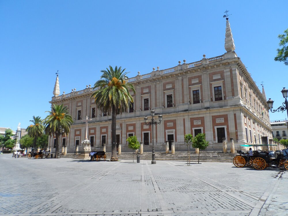 Fachada do edifício do Arquivo geral das Índias, em Sevilha, com paredes em tons claros e com muitas janelas.