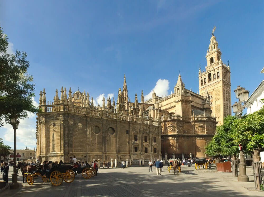 Praça com carruagens, pessoas, árvores, a Catedral de Sevilha e a Torre La Giralda.