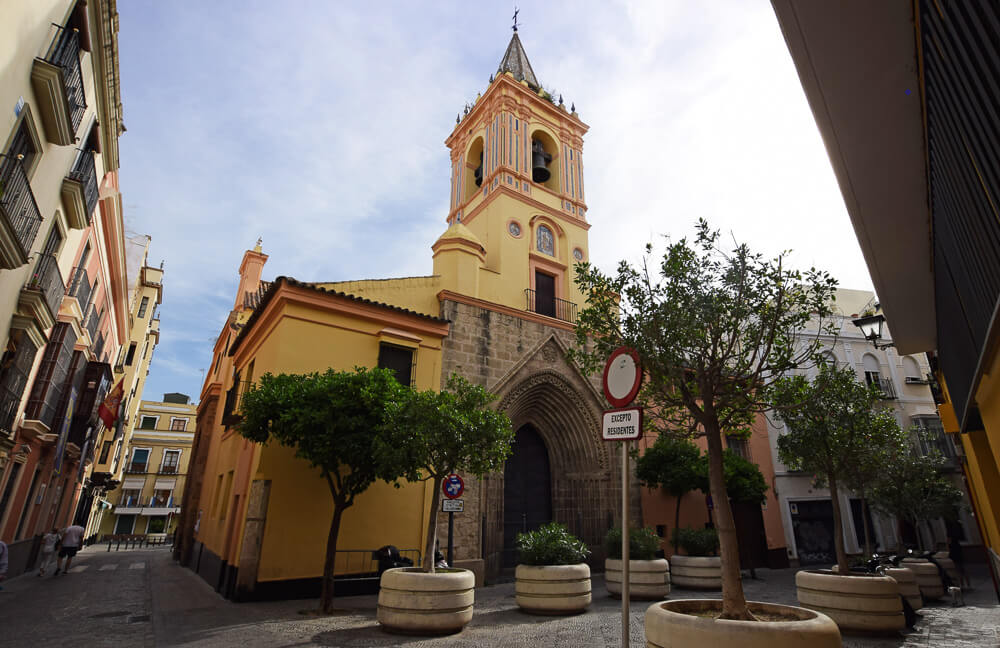 Entrada da Igreja de San Isidoro, com fachada amarela, algumas árvores e prédios baixos nas redondezas.