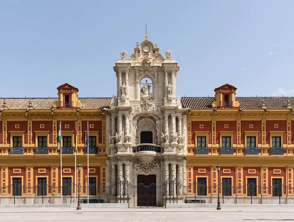Fachada do Palacio de San Telmo, com paredes vermelhas, laranja e esculturas no centro.