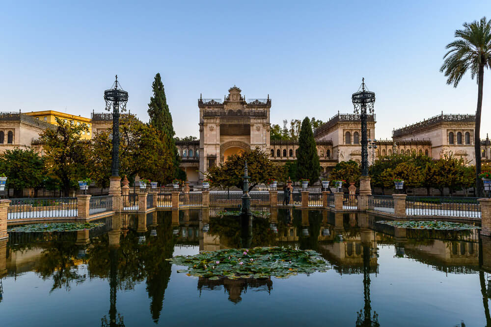 Fachada do Museu Arqueológico de Sevilha, com várias árvores e um lago em frente.