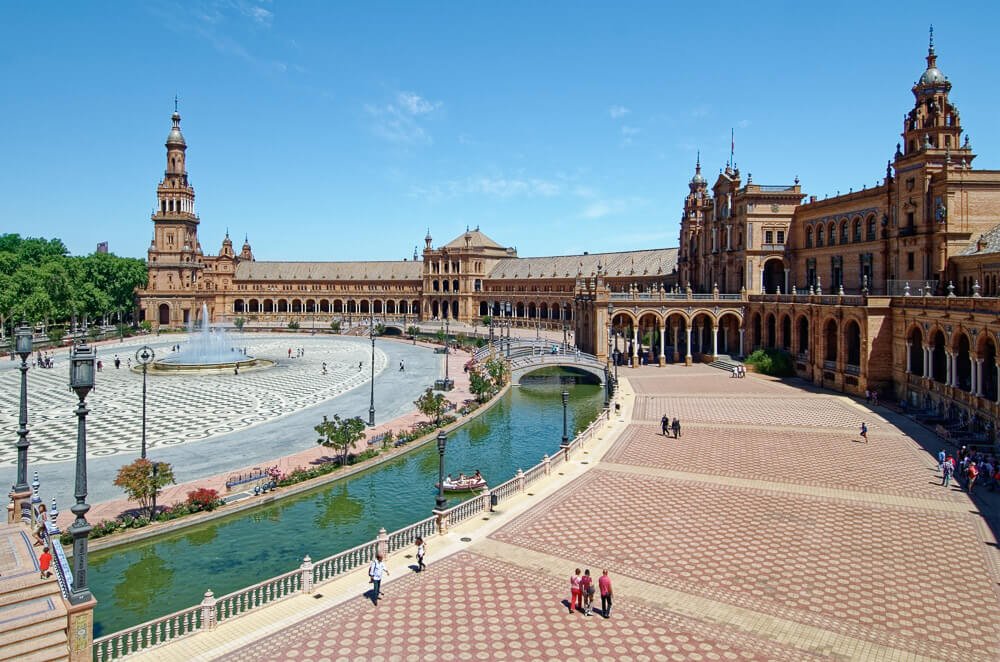 Plaza de España em um dia de sol, com fachada semicircular, muitos arcos e um pequeno canal de águas verdes.