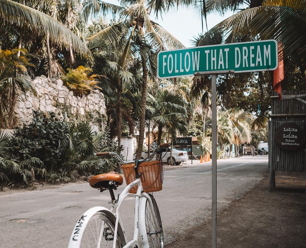 Placa Follow That Dream, com uma bicicleta ao lado, em uma estraga estreita de Tulum.