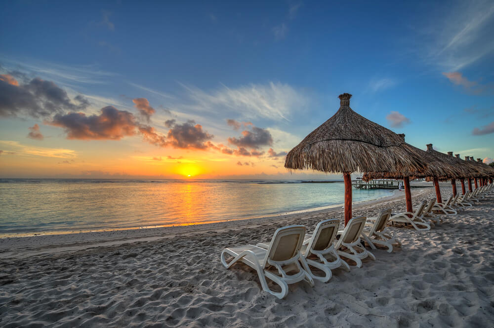 Playa Akumal com várias espreguiçadeiras, toldos de palha e o pôr do sol refletindo no mar.