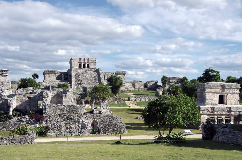 Zona arqueológica de Tulum com várias ruínas Maias, árvores e um gramado.