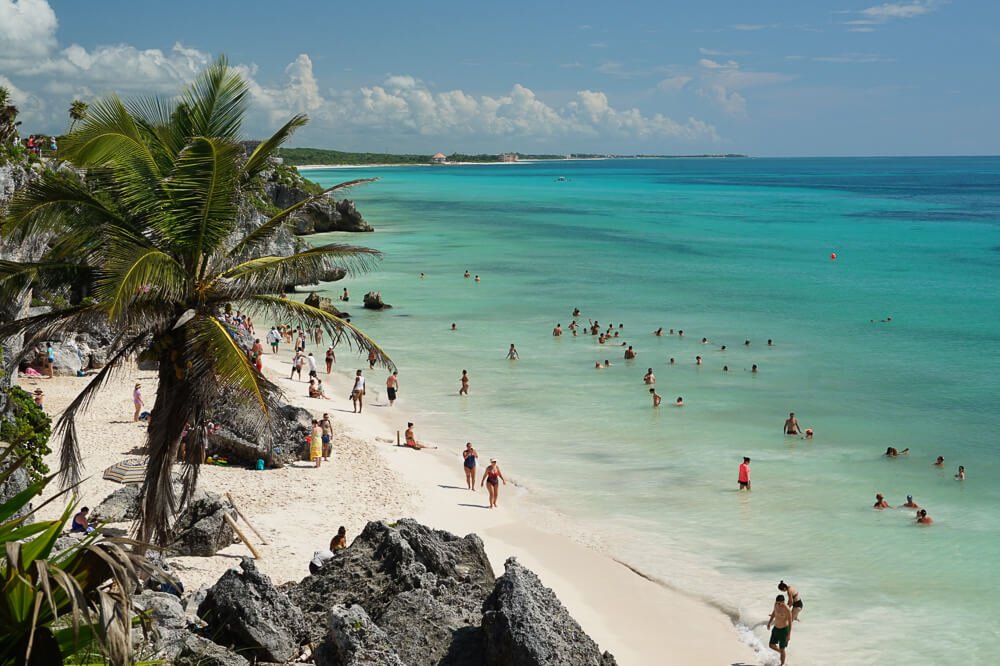 Playa de Tulum, com coqueiros, pedras, águas cristalinas e vários banhistas.