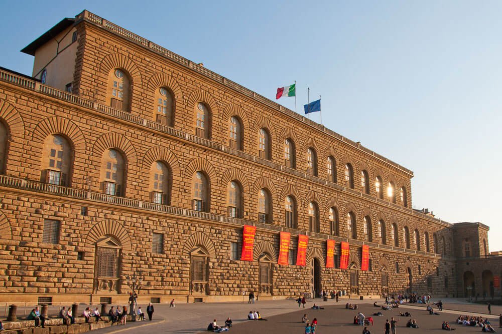 Fachada do Palazzo Pitti com paredes de tijolos marrons e várias pessoas no pátio em frente ao edifício.