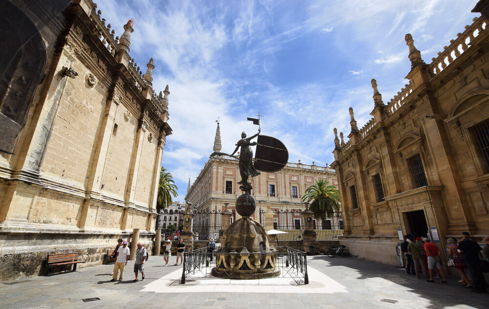 Entrada da Catedral de Sevilha, com um portão gradeado, pessoas e a estátua Giraldillo no centro.