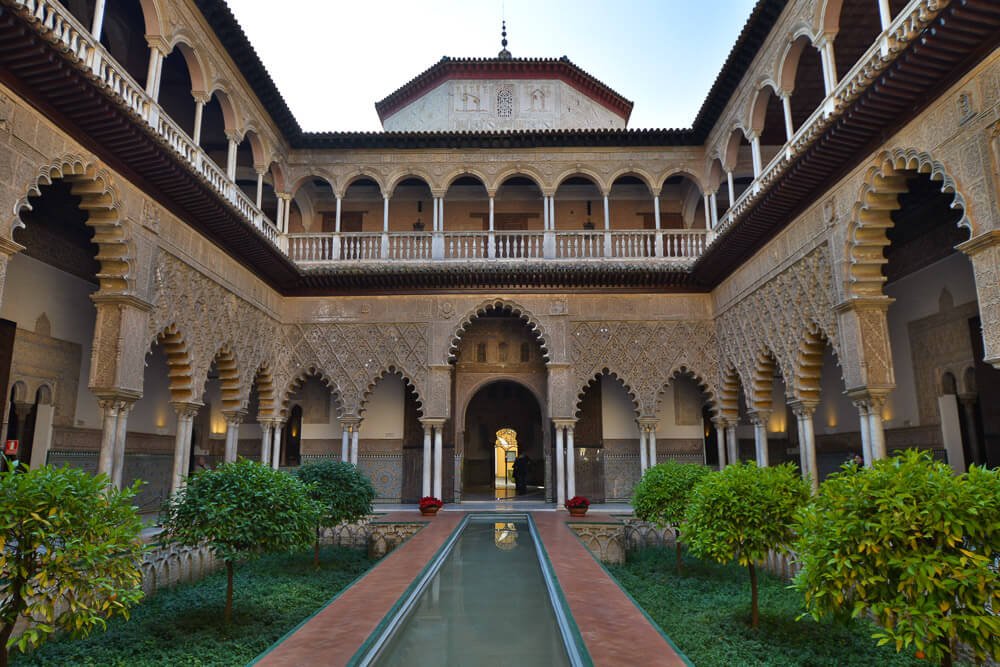 Interior do Alcázar de Sevilha, com arcos, um pequeno jardim e um lago artificial.