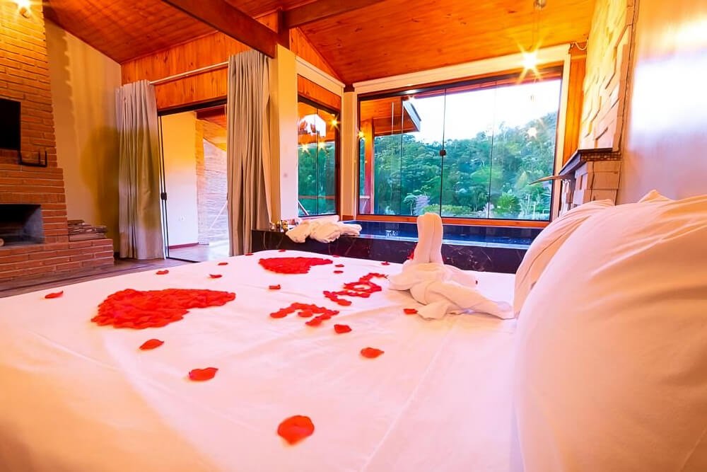Quarto da pousada Chalé Luiz com cama, lareira e banheira de hidromassagem com vista para a natureza.
