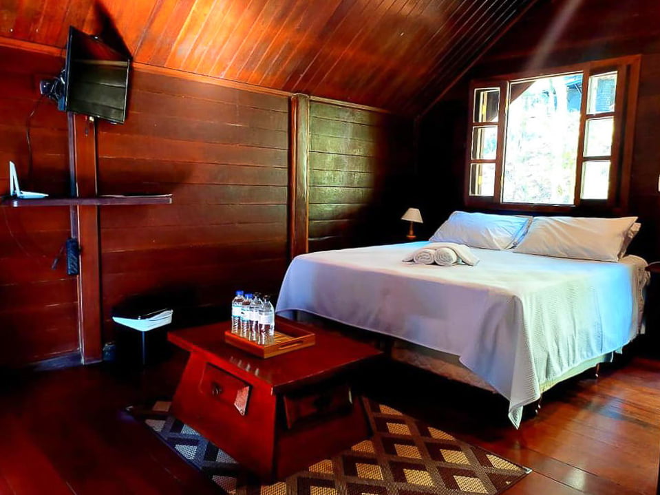 Quarto da pousada Pássaro de Fogo, com cama, TV, paredes de madeira e mesa de centro.
