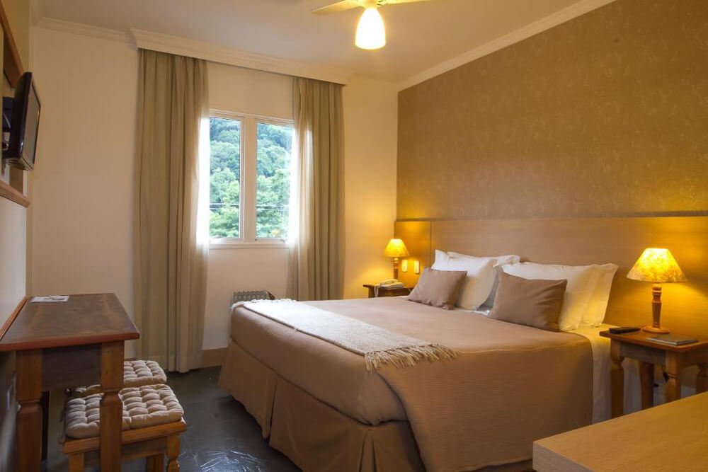 Quarto da Pousada Il Villaggio com cama, TV, iluminação aconchegante, móveis de madeira e janela.