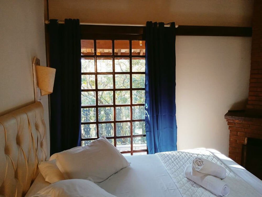Quarto da Pousada Le Lieu com cama, janela, cortina e lareira.
