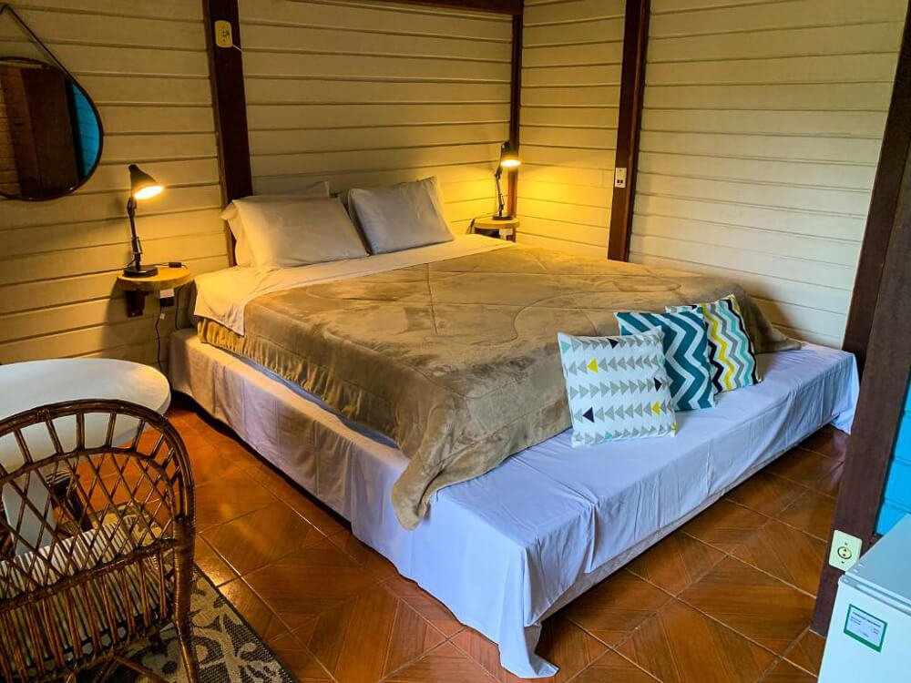 Quarto da pousada Recanto das Águas, com paredes de madeira clara, cama, espelho, mesa e cadeira.