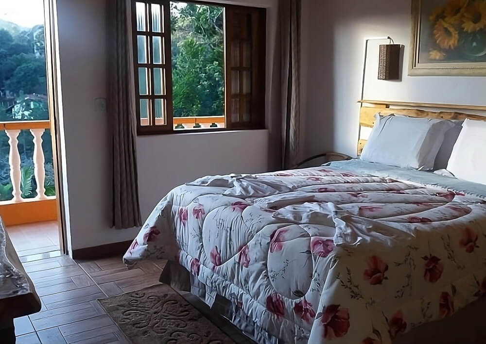 Quarto da Pousada Pinhalense com cama, quadro com pintura de girassóis, janela e varanda com vista para a natureza.