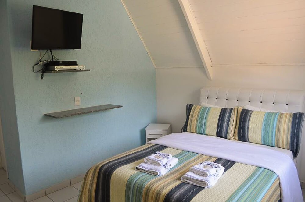 Quarto da Pousada Recanto das Orquídeas, com cama, prateleira e TV.