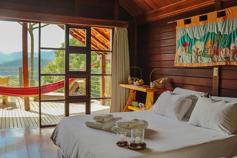 Quarto da Pousada Vento Verde com cama, móvel com frutas, paredes de madeira, varanda com rede e vista para a serra.