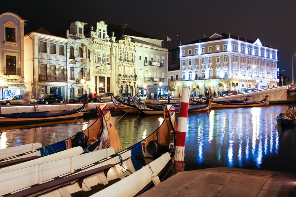 Ria de Aveiro à noite, com barcos moliceiros e prédios iluminados.