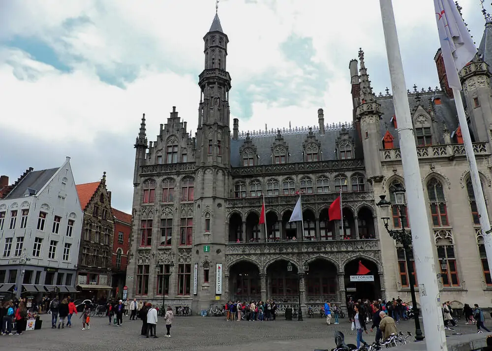 Fachada do Museu Bruges Historium, em Bruges, com vários arcos, uma torre e paredes cinza.