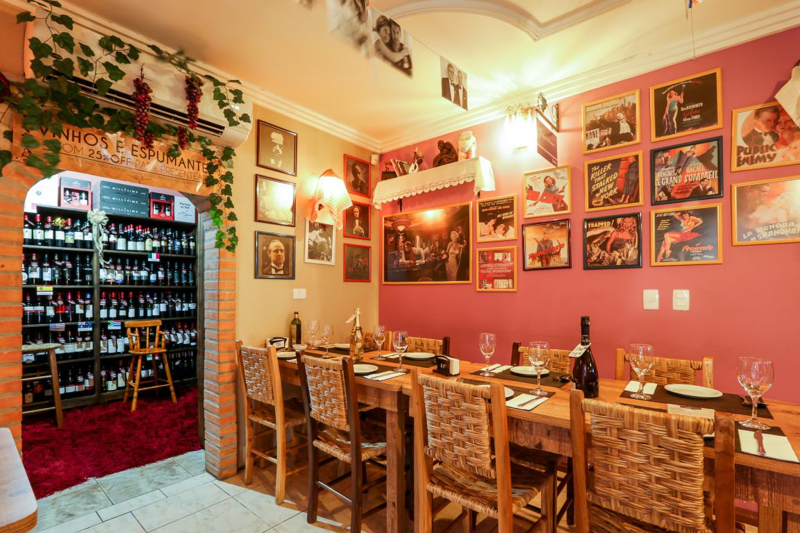 Interior do restaurante La Mafia Trattoria em Foz do Iguaçu. As paredes são decoradas com quadros. À direita, há uma mesa de madeira retangular com 8 cadeiras. À esquerda, há uma porta de entrada para uma adega cheio de vinhos