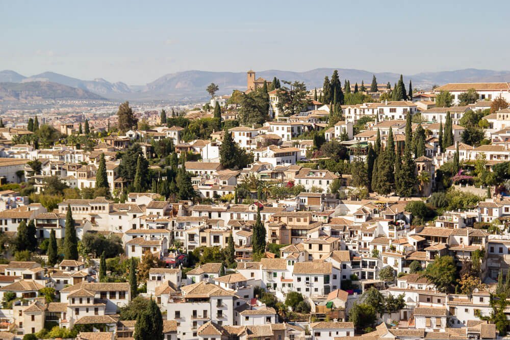 Casas caiadas de Albaicín vistas de cima, com várias árvores italianas e montes ao fundo.