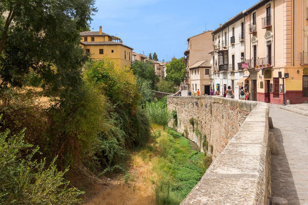 Rua Carrera del Darro com uma pequena vegetação à esquerda e prédios baixos à direita.