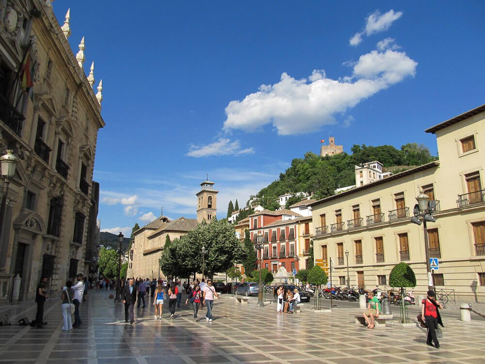 Plaza Nueva, com edifícios cheios de janelas, igreja, árvores e pessoas caminhando.