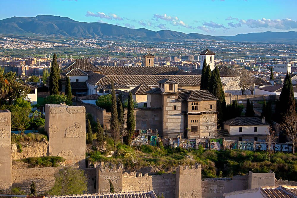 Vista de um local alto para o Palácio de Dar al-Horra, com muitas árvores ao redor e montanhas ao fundo.