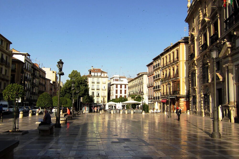 Plaza Nueva iluminada pela luz do sol, com árvores, luminárias públicas e prédios por todos os lados.