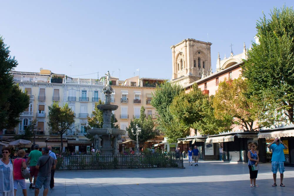Praça Bib-Rambla com prédios, árvores, uma fonte e pessoas caminhando.