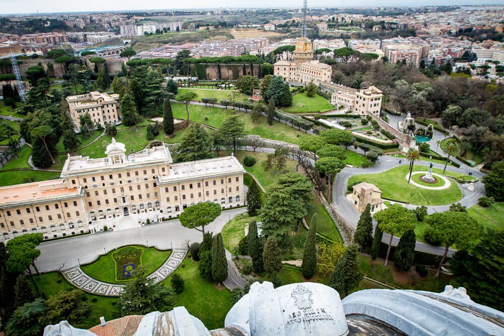 Panorama aéreo dos Jardins do Vaticano, com alguns prédios claros, gramados e árvores.