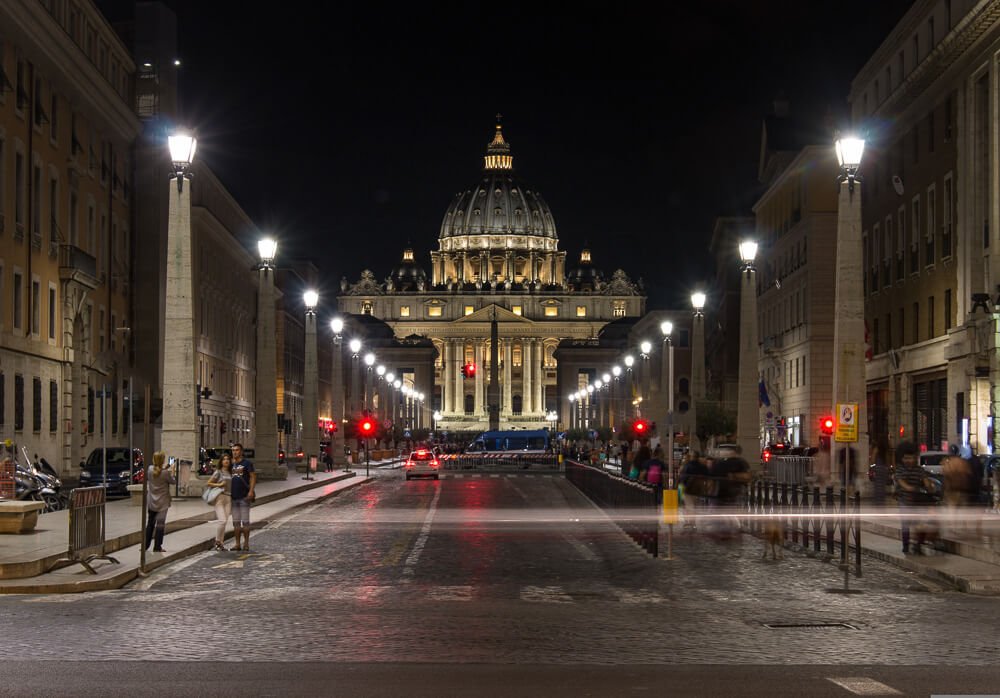 Via della Conciliazione à noite, com alguns pedestres e a Basílica de São Pedro no centro.