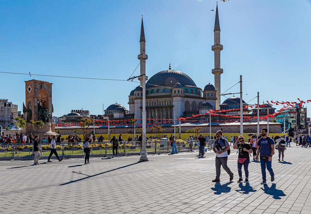 Praça Taksim com muitos visitantes e uma mesquita ao fundo.