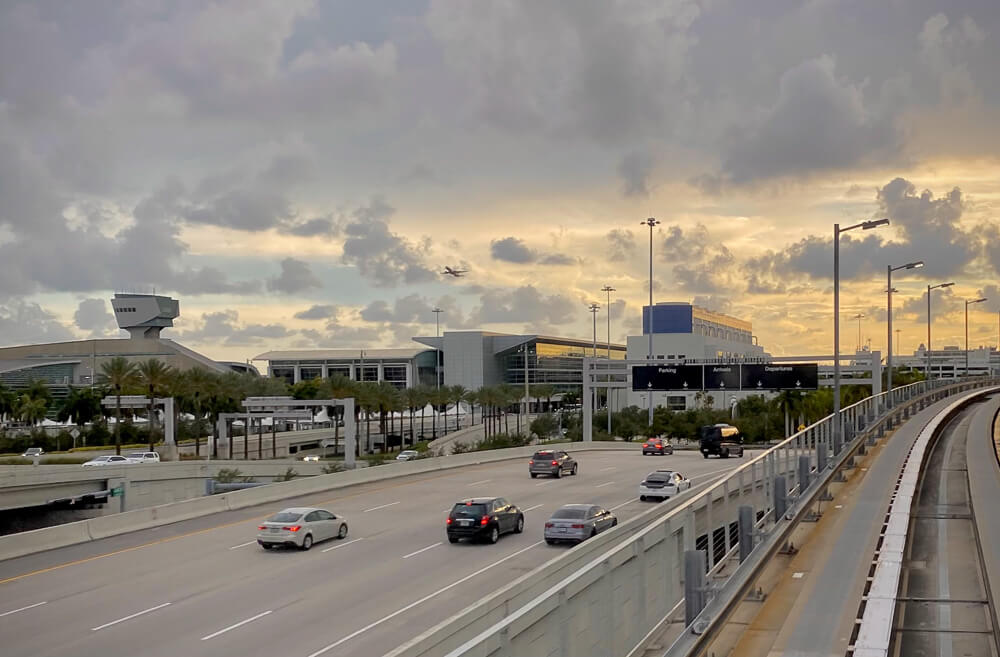 Vias nas proximidades do Aeroporto Internacional de Miami, com vários carros e o sol poente ao fundo.