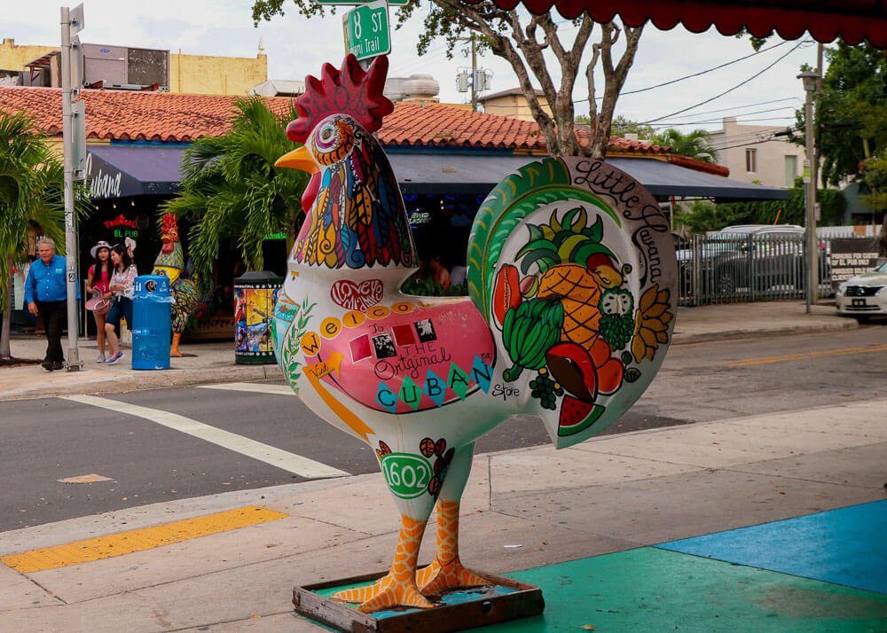Rua Calle Ocho, em Little Havana, com escultura de galo colorido no centro e vários estabelecimentos ao fundo.