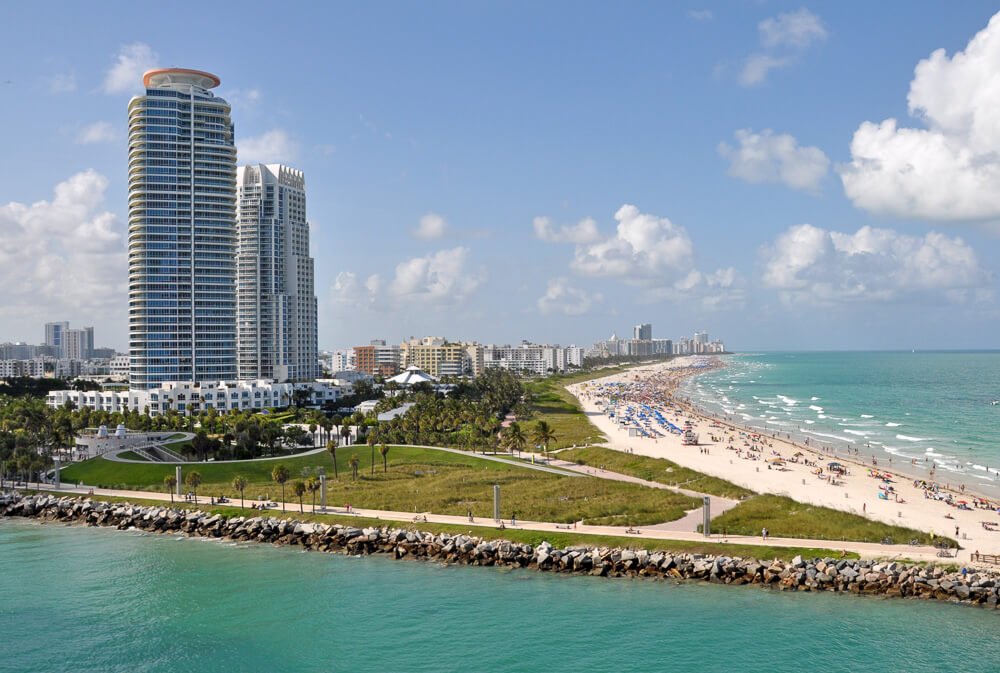 South Beach, em Miami, com várias árvores, praia com areia clara, mar de águas cristalinas e alguns prédios de frente ao mar.
