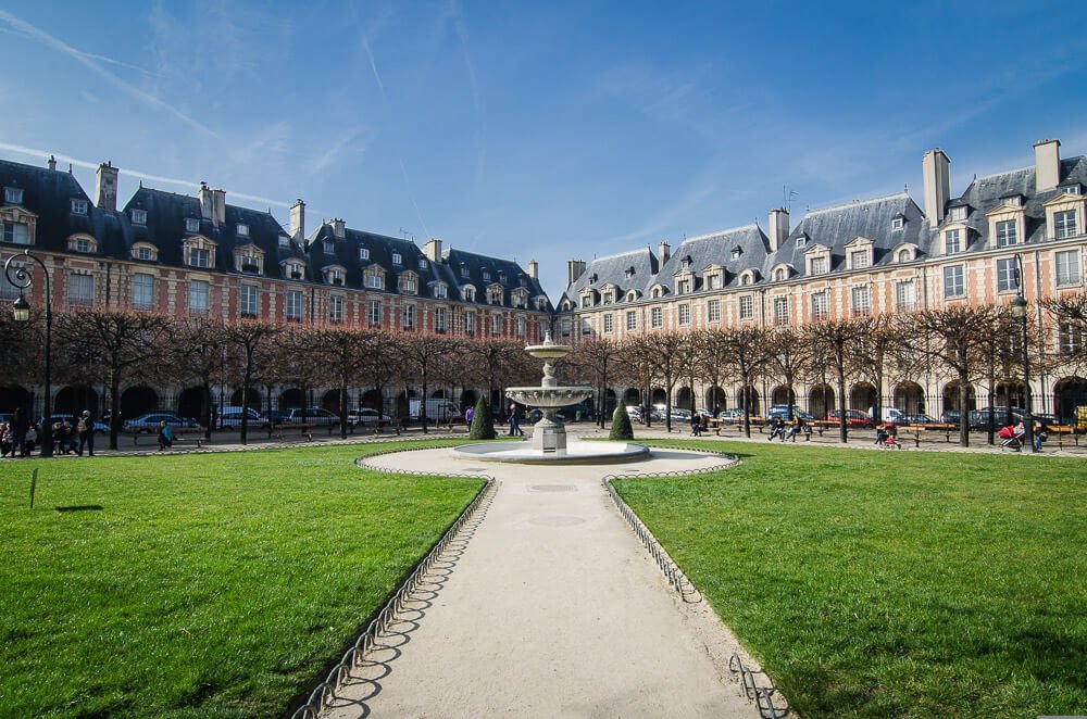 Place des Vosges com um gramado, uma fonte, várias árvores e prédios ao redor.