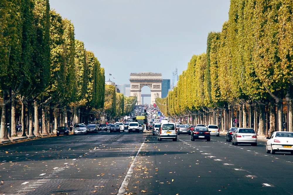 Champs-Élysées com muitos carros, árvores nas laterais e o Arco do Triunfo ao final da rua.