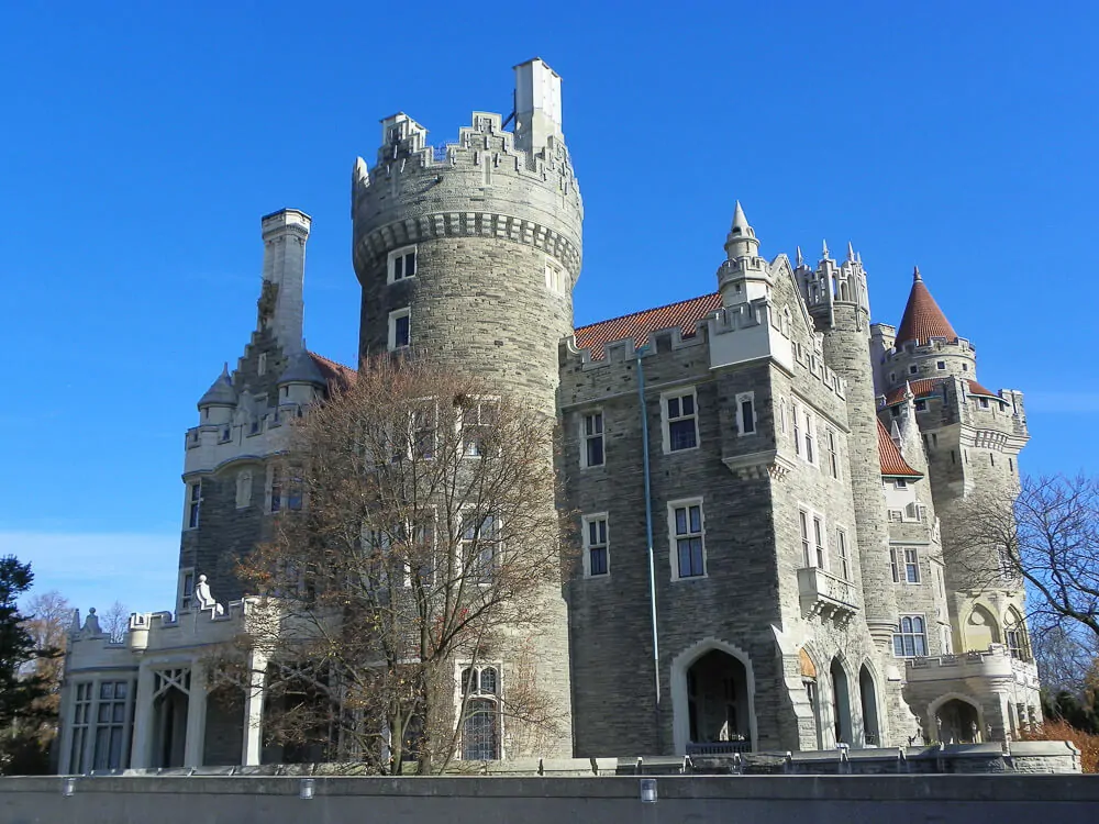 Fachada da casa Loma, com arquitetura neo-romântica, paredes de pedras e algumas árvores em frente.
