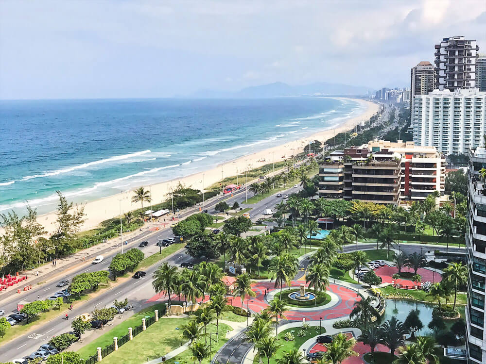 Barra da Tijuca vista de cima, com um mar agitado, avenidas costeiros e prédios à beira-mar.
