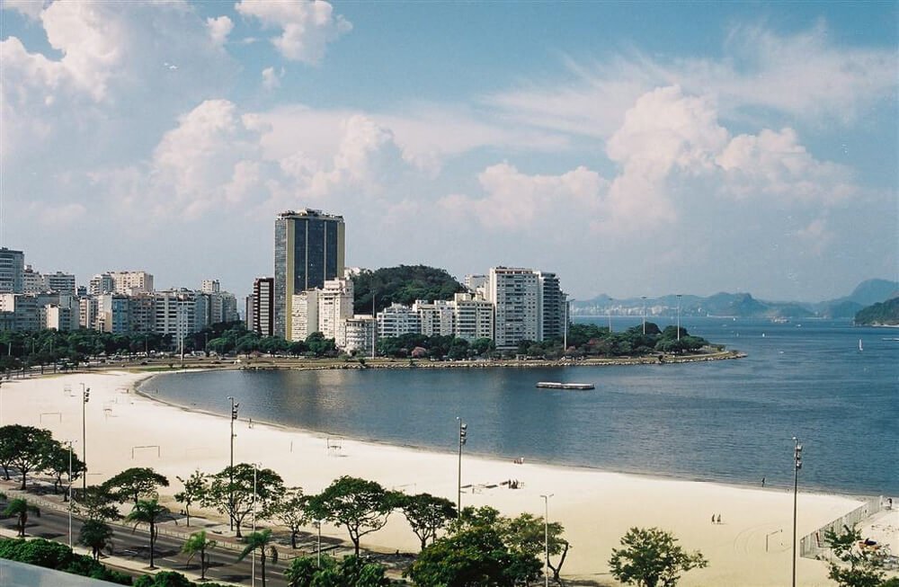 Praia de Botafogo, na Zona Sul do Rio de Janeiro, com faixa de areia extensa, mar de águas calmas e escuras, prédios à beira-mar e várias árvores.