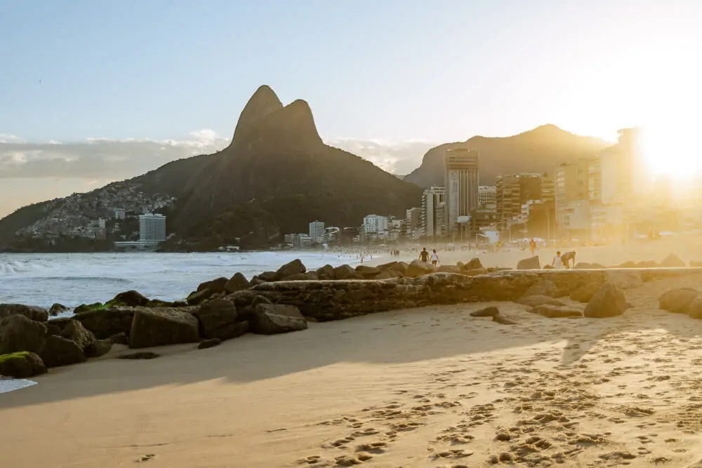 Tarde ensolarada na Praia de Ipanema, com mar agitado, grande faixa de areia, algumas pedras e o Morro Dois Irmãos ao fundo.
