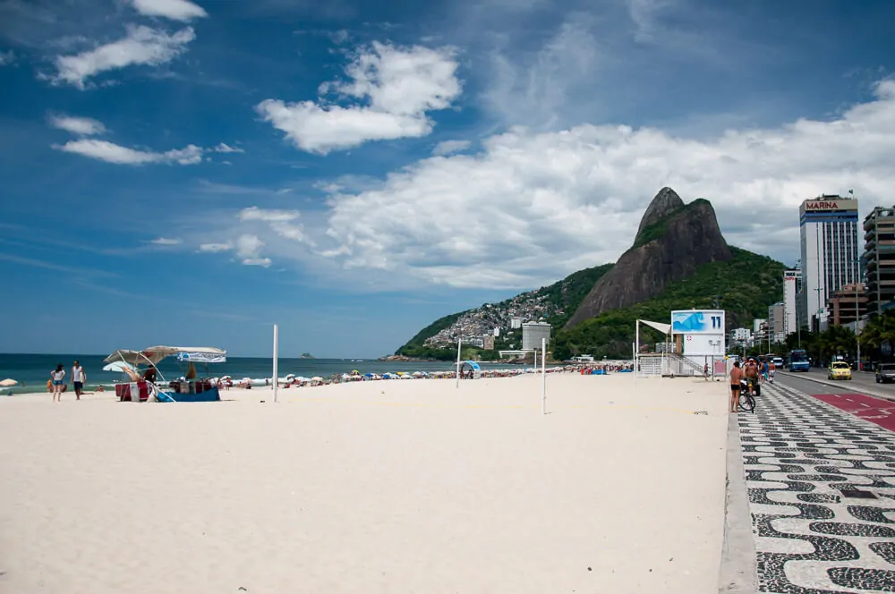 Praia do Leblon, com uma extensa faixa de areia, mar calmo, prédios à beira-mar e o morro Dois Irmãos ao fundo.