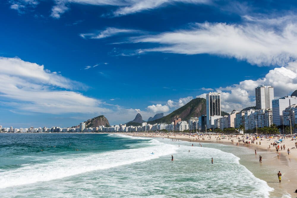 Praia do Leme, com águas esverdeadas, muitos banhistas, algumas ondas e prédios em frente ao mar.