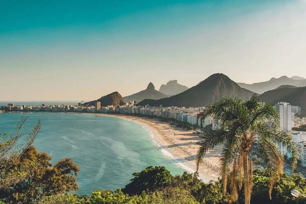 Praia de Copacabana vista do alto, com águas cristalinas, prédios em frente ao mar e morros ao fundo.
