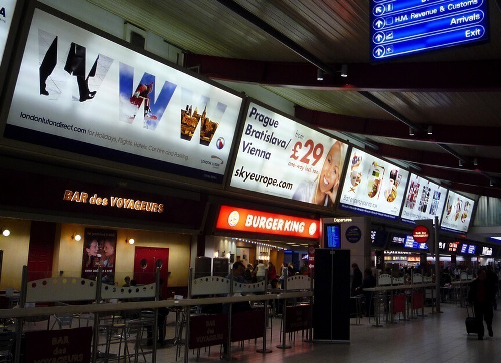 O interior do Aeroporto de Luton, com estabelecimentos comerciais e publicidade.