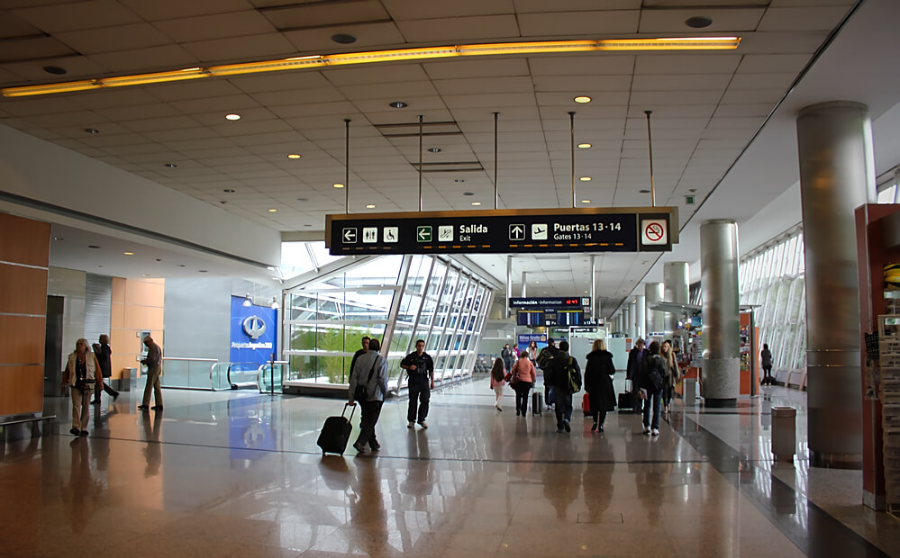 Visão interna do Aeroporto Jorge Newberry, várias pessoas estão andando e carregando bagagens.